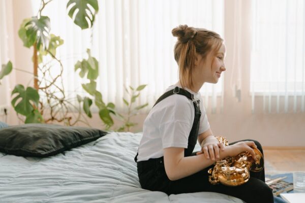 A young girl sitting on bed indoors, holding a saxophone, surrounded by plants.
