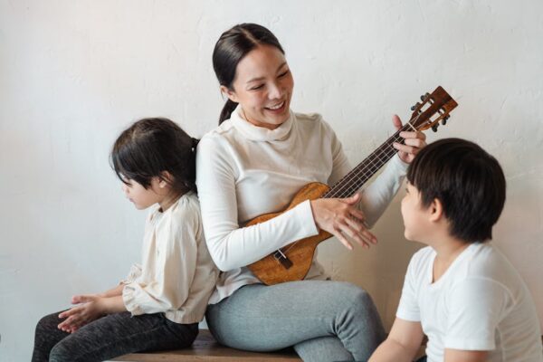 Happy musician sitting with crossed legs while playing traditional Hawaiian musical instrument for happy unrecognizable ethnic son and pensive anonymous daughter sitting on wooden bench at home