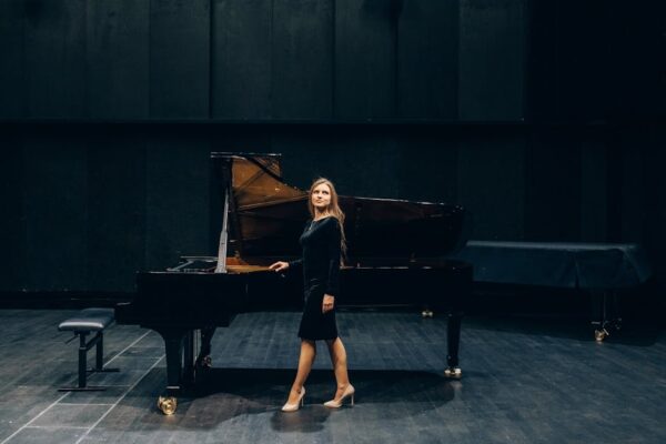 An elegant woman in a black dress standing next to a grand piano on stage indoors.