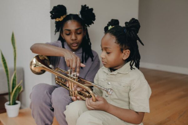Two children sitting indoors, one teaching the other to play a brass trumpet, creating a warm family moment.