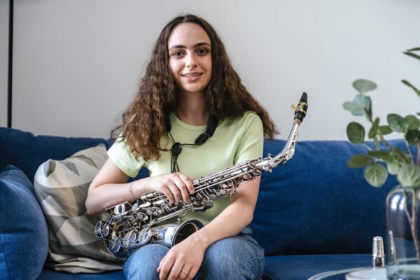 A young woman sitting on a couch holding a saxophone, smiling indoors.