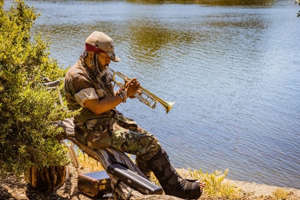 A man in a camo uniform playing a trumpet by the riverside. Calm and serene outdoor setting.