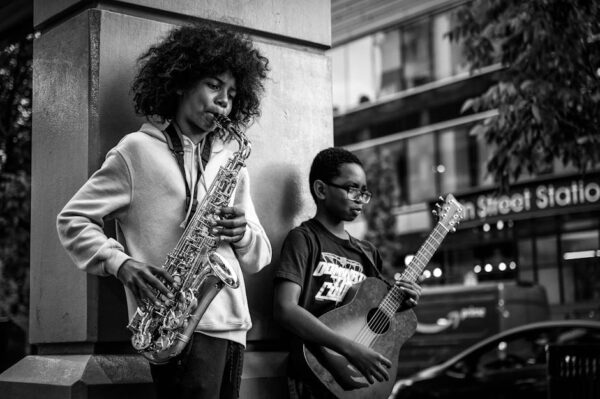 Black and white of African American guy with curly hair standing and playing saxophone with friend guitarist on city street in daytime