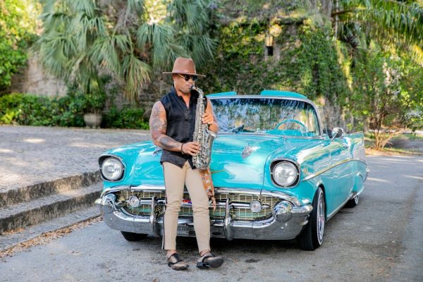 A stylish saxophonist plays music beside a vintage turquoise car on a sunny day.