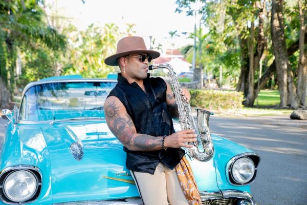 A trendy musician plays saxophone leaning on vintage blue car outdoors.