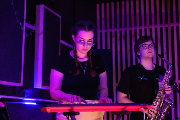 Musicians performing with a keyboard and saxophone under dramatic lighting.