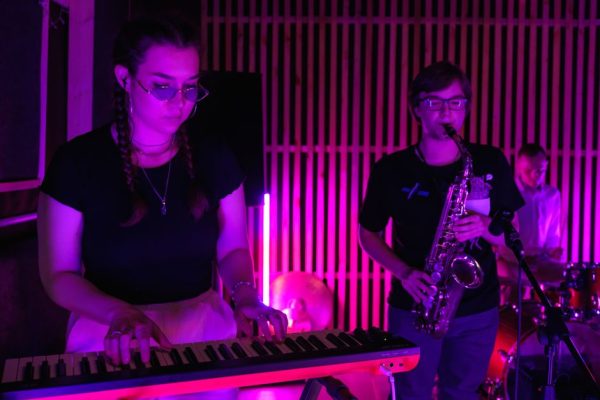 Energetic live performance with keyboard, saxophone, and drums under vibrant lighting.
